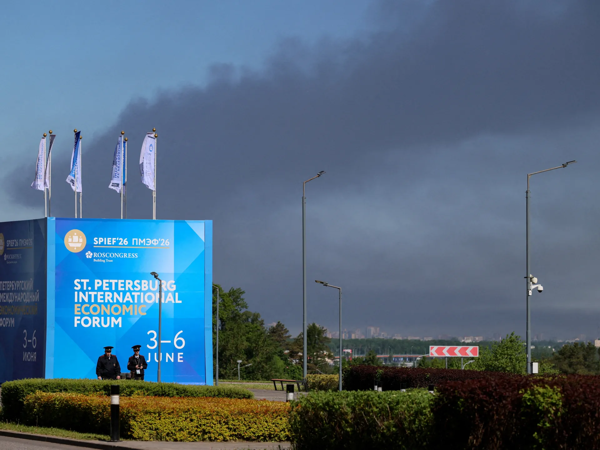 Police officers stand in front of a banner and flags with the logo of the Saint Petersburg International Economic Forum (SPIEF) as heavy smoke billows in the background after Ukrainian drones hit infrastructure, in St Petersburg, Russia, on June 3, 2026 [Reuters]