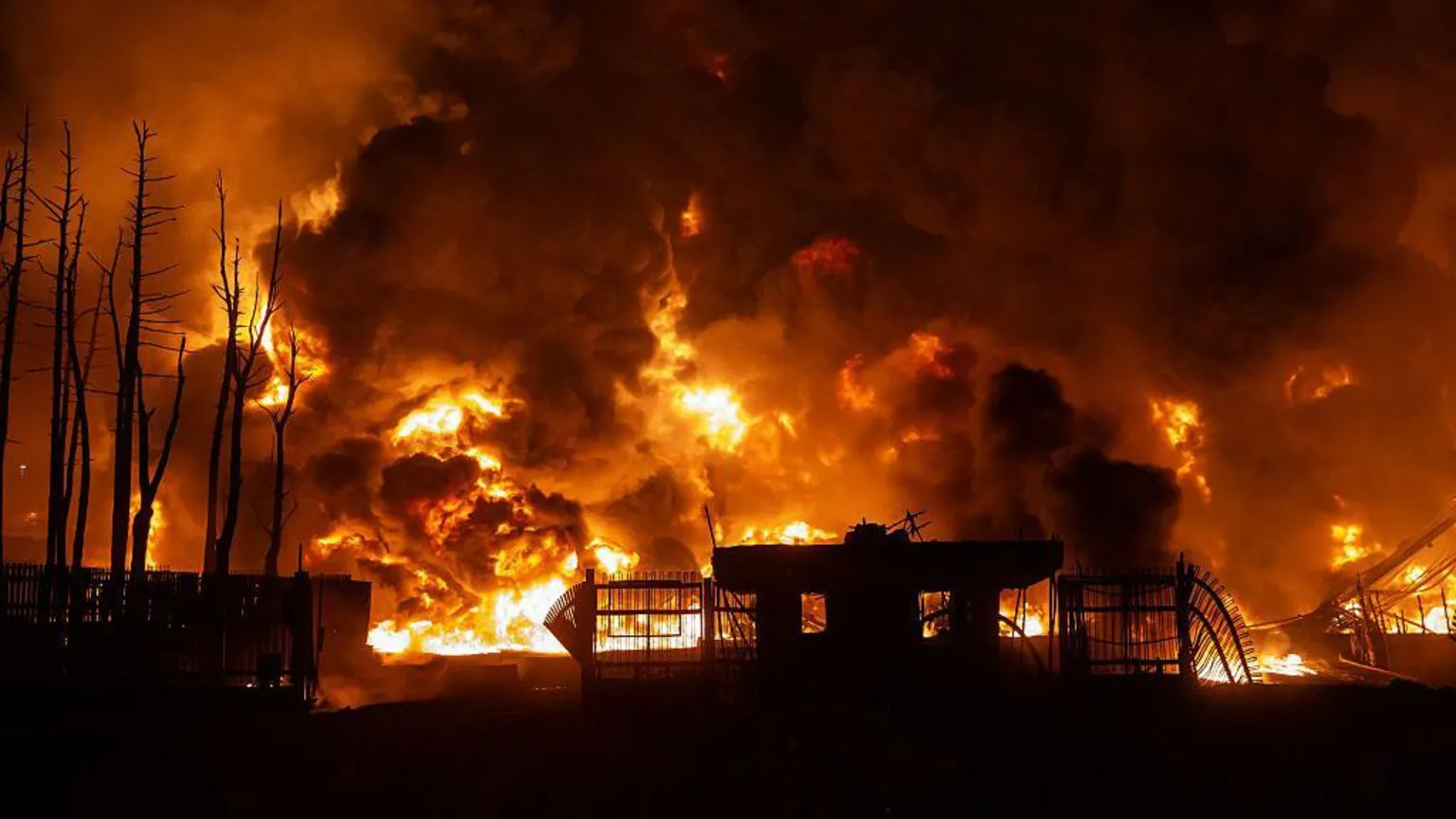Anadolu via Getty Images A large fire burns at an oil depot in Iran at night. Flames and thick plumes of dark smoke tower over the outline of a small building.